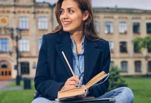 Estudante sorridente sentada ao ar livre com um caderno e um lápis, em frente a um prédio universitário, representando exemplos de bolsas de estudo disponíveis.